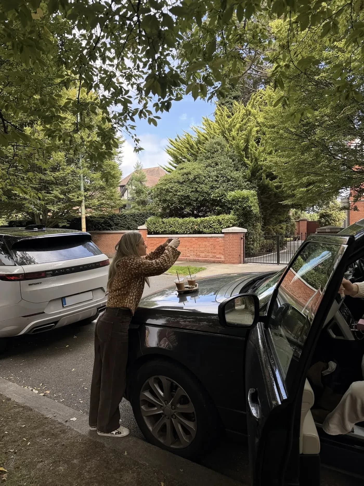 Rebeccah leaning over a car bonnet to photograph two iced coffees on a leafy Dublin street